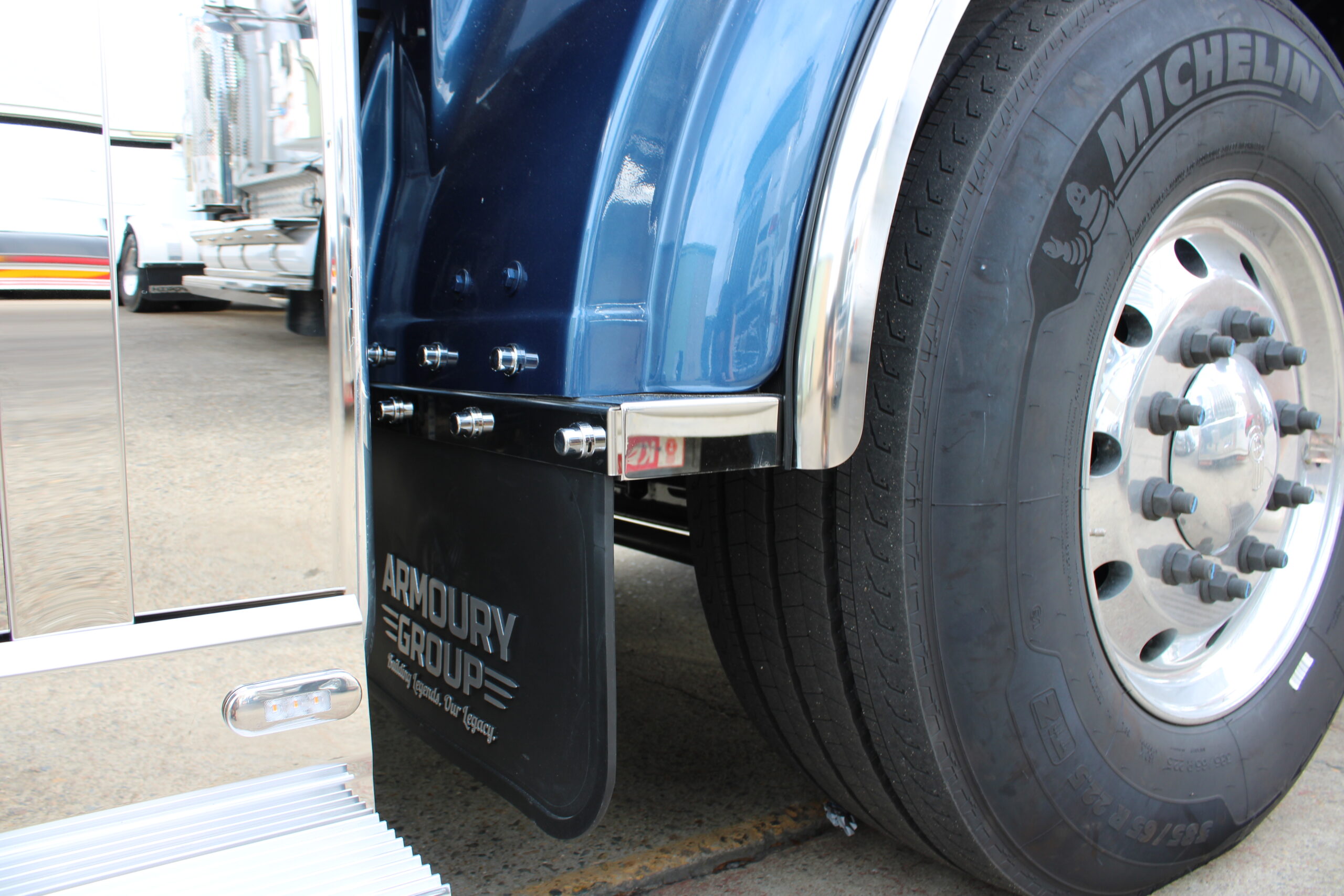 Close-up of a large trucks rear wheel and tire, showing the blue fender, Michelin tire, chrome accents, and a black mud flap labeled ARMOURY GROUP with metal fasteners visible.