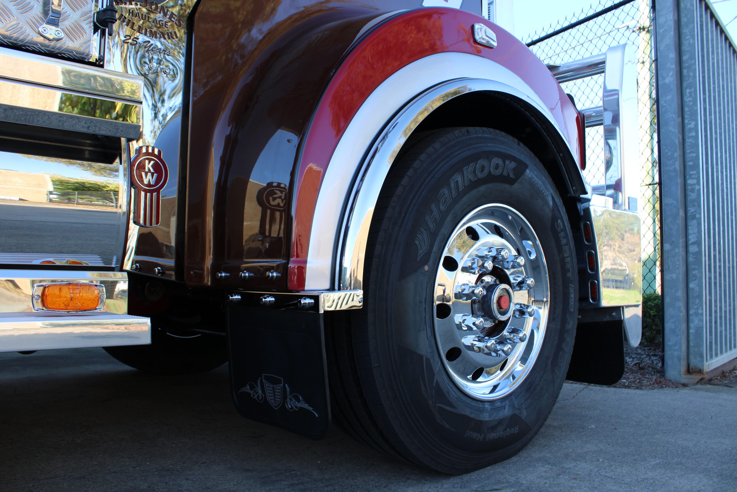 Close-up of the rear wheel and fender of a shiny maroon semi-truck, showing chrome details, a Kenworth emblem, Hankook tire, and a black mud flap with a stylized emblem, parked near a chain-link fence.
