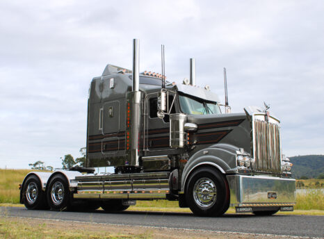 A shiny, dark gray semi truck with chrome accents and large exhaust stacks is parked on a country road with grassy fields and hills in the background under a cloudy sky.