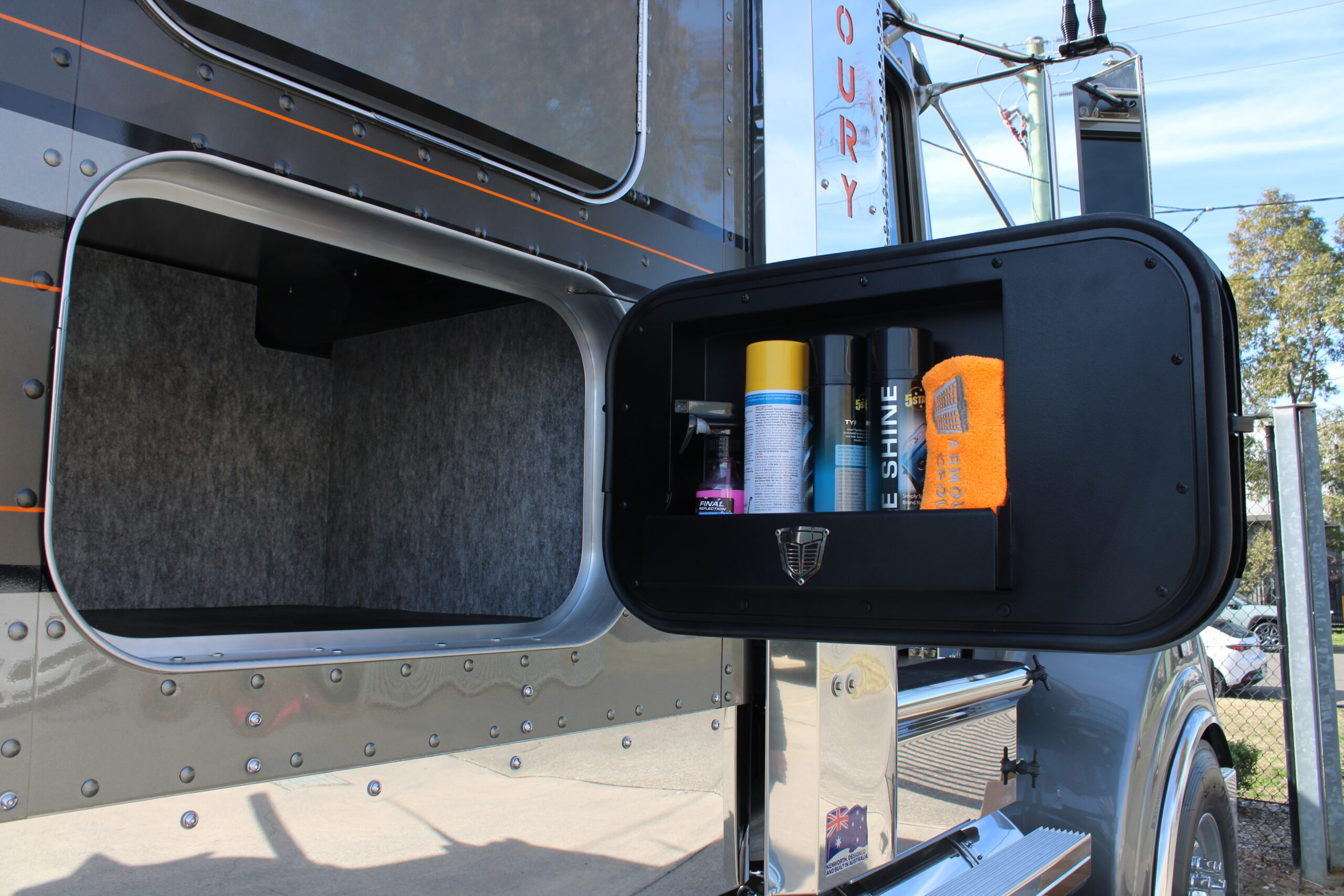 A storage compartment on the side of a truck, opened to reveal cleaning supplies including spray cans, a towel, and a can labeled SHINE. The truck’s metallic exterior is visible in the background.