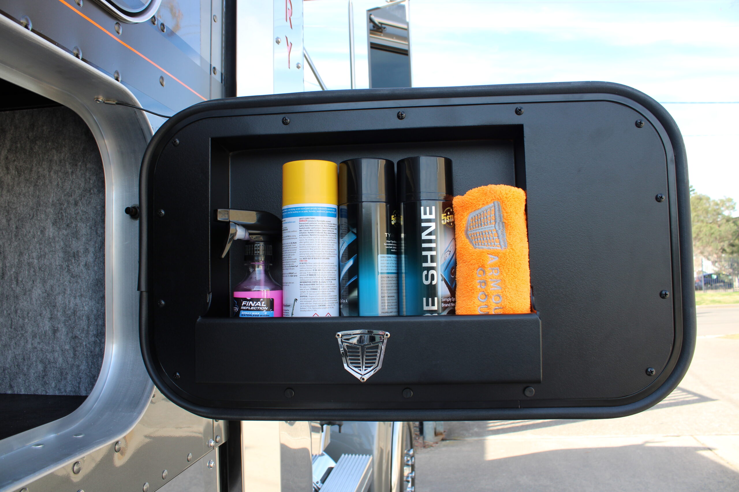 A close-up of an open truck storage compartment door holding cleaning supplies: three spray cans, a spray bottle, and an orange microfiber towel, parked outdoors on a sunny day.