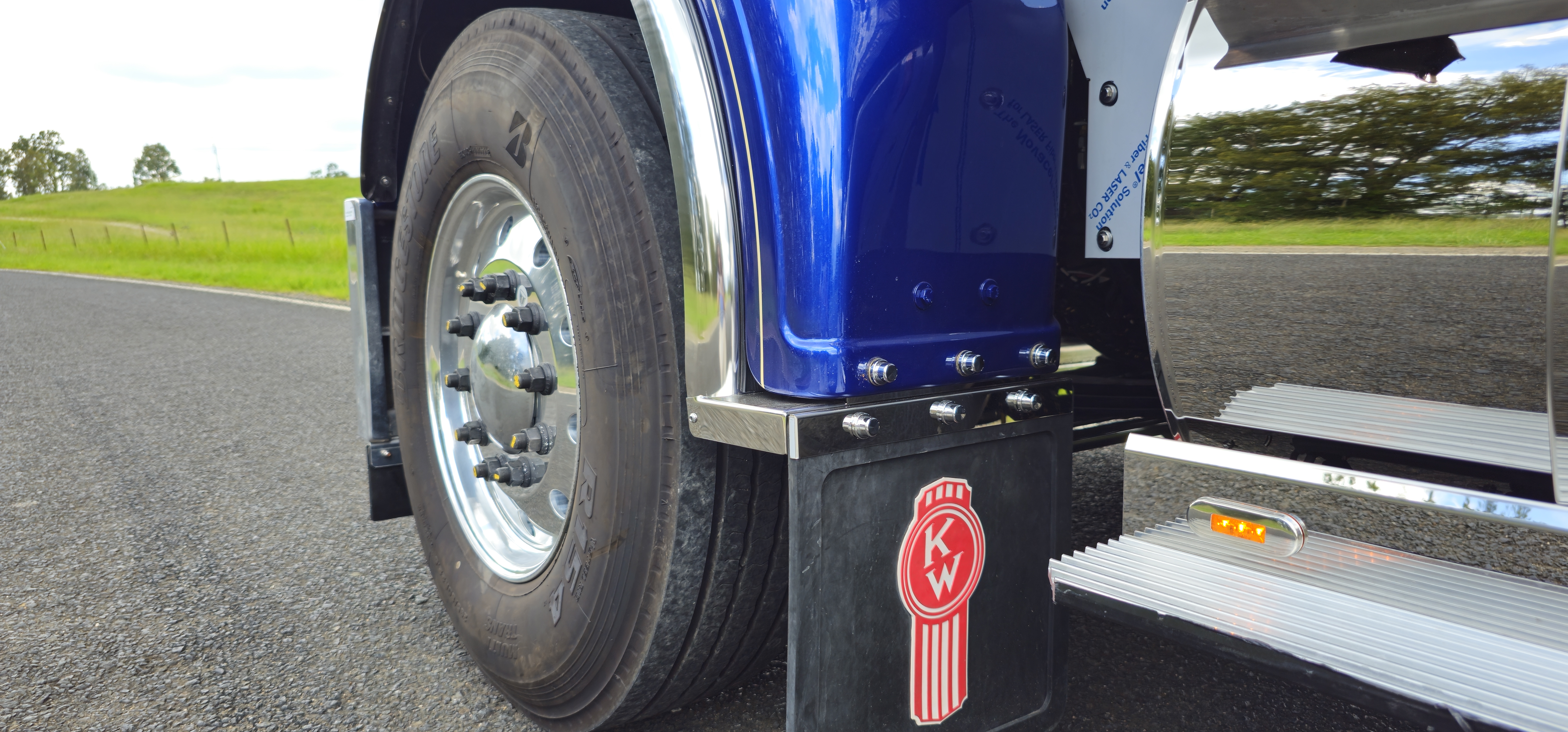 Close-up of a blue trucks rear wheel and mud flap with a Kenworth logo, parked on an asphalt road with green grass and trees in the background.